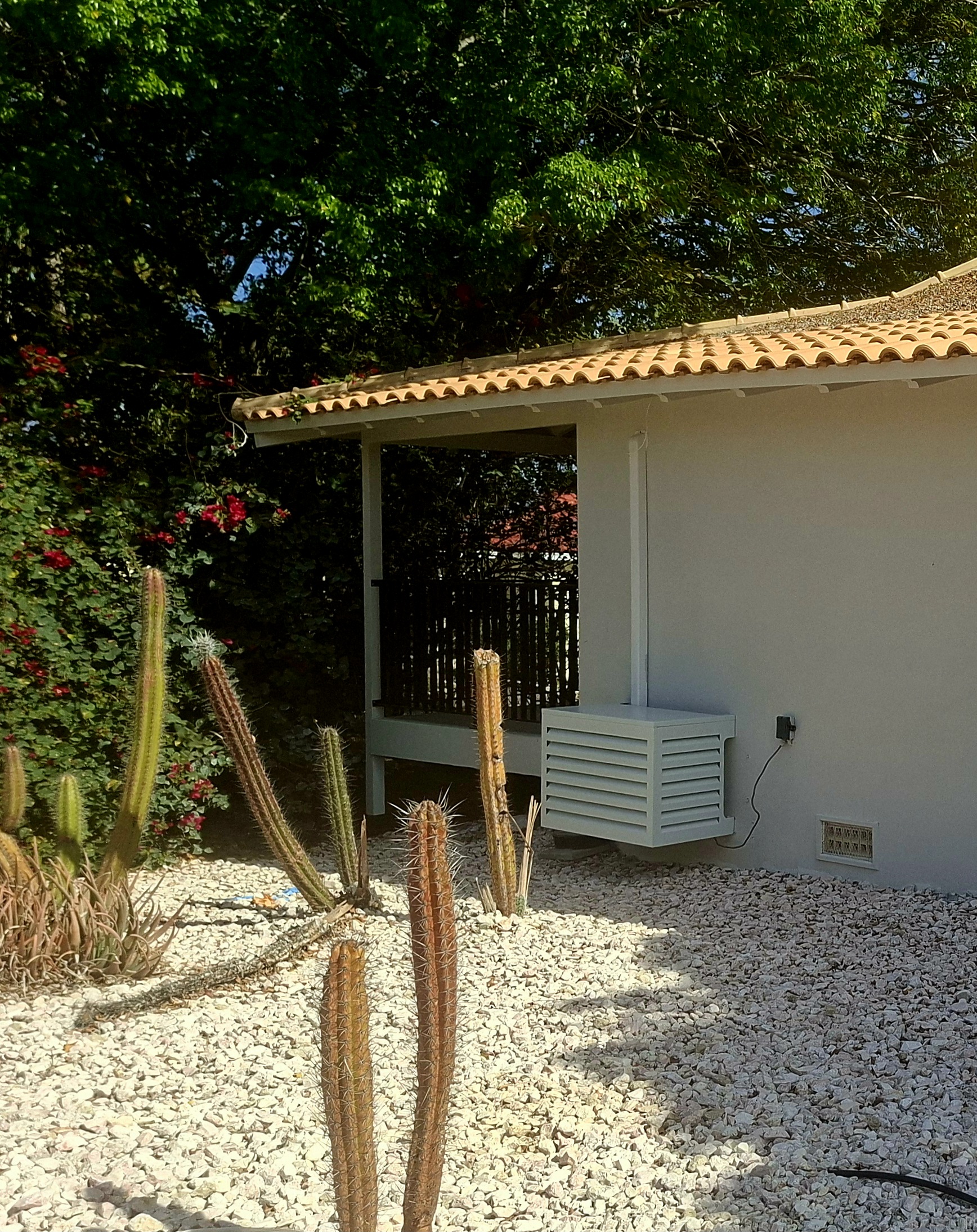 Aluminum AC cover mounted on a Curacao home beside a gravel garden with cacti and bougainvillea
